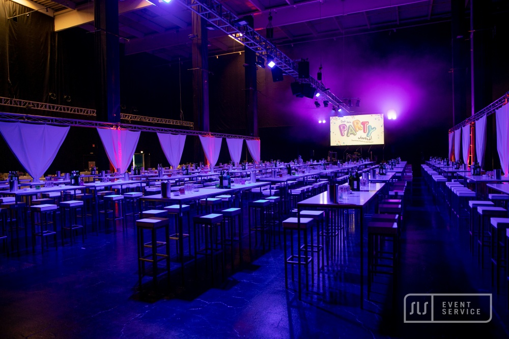 Eventhalle mit langen Stehtischen, Sitzplätzen, Bühne und violetter Beleuchtung