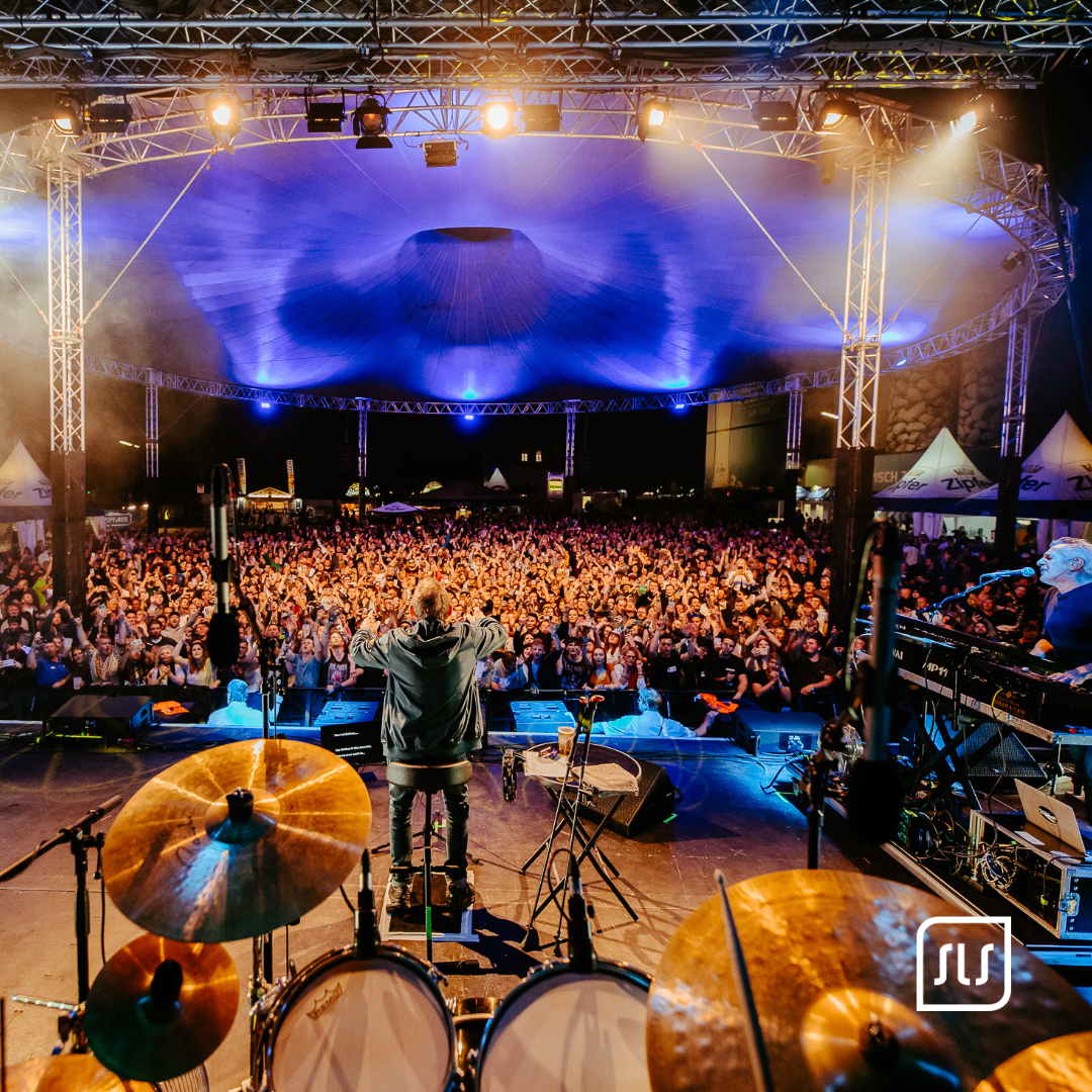 Blick von der ZipfAir-Bühne über Schlagzeug und Musiker auf ein dichtes Konzertpublikum unter blau beleuchtetem Zeltdach