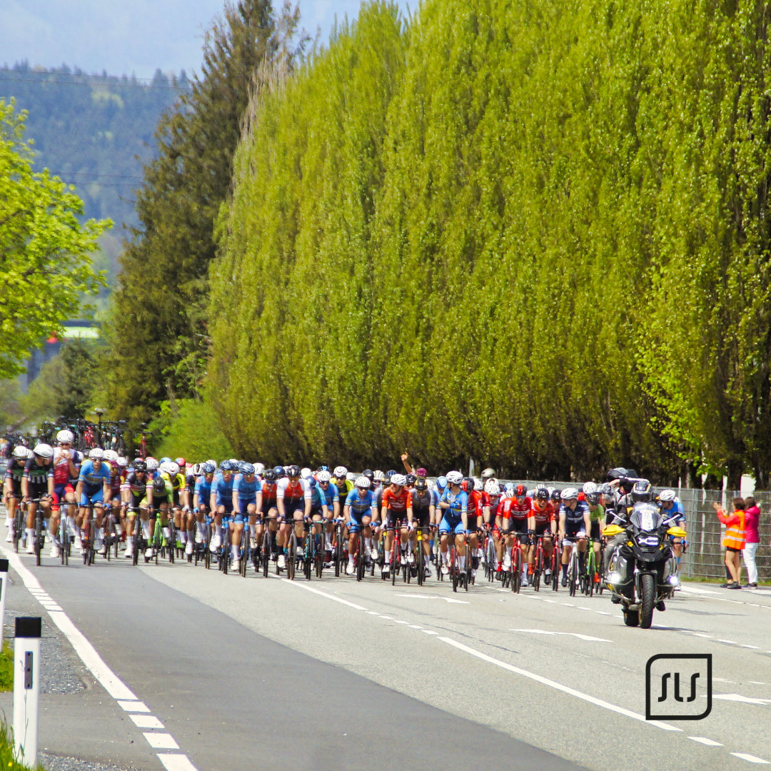 Großes Peloton beim 12. GP Vorarlberg fährt hinter einem Begleitmotorrad auf einer von Bäumen gesäumten Straße