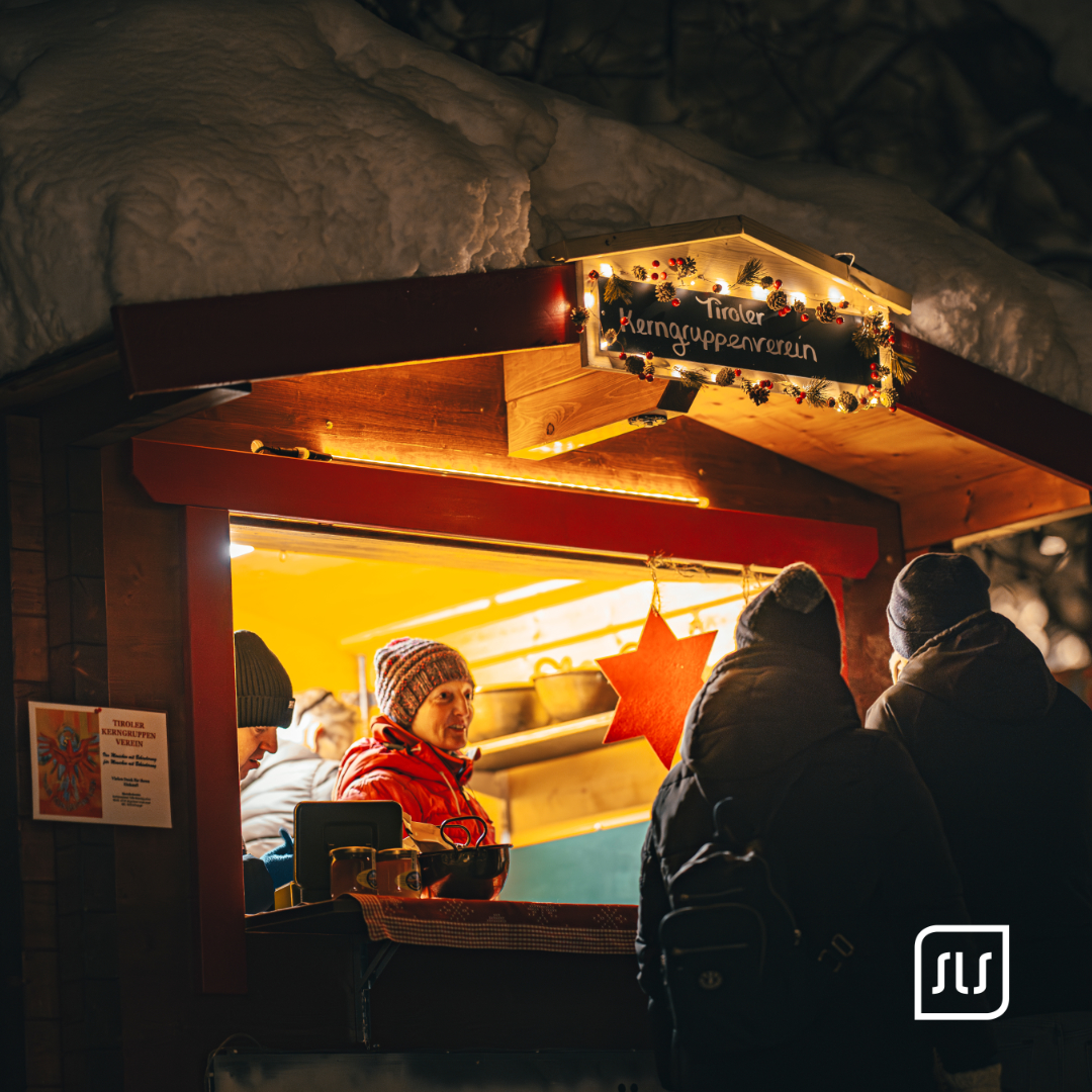 Gäste stehen an einem warm beleuchteten Holzstand mit Schneedecke beim Adventzauber in St. Anton