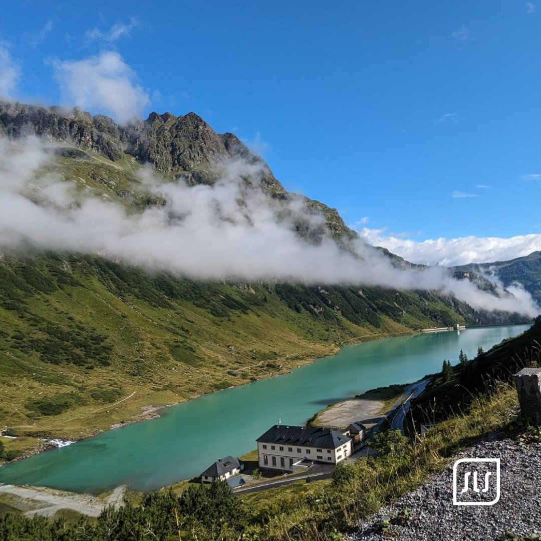 Türkisfarbener Bergsee unter felsigen Gipfeln und tiefen Wolken entlang der Arlberg-Giro-Strecke