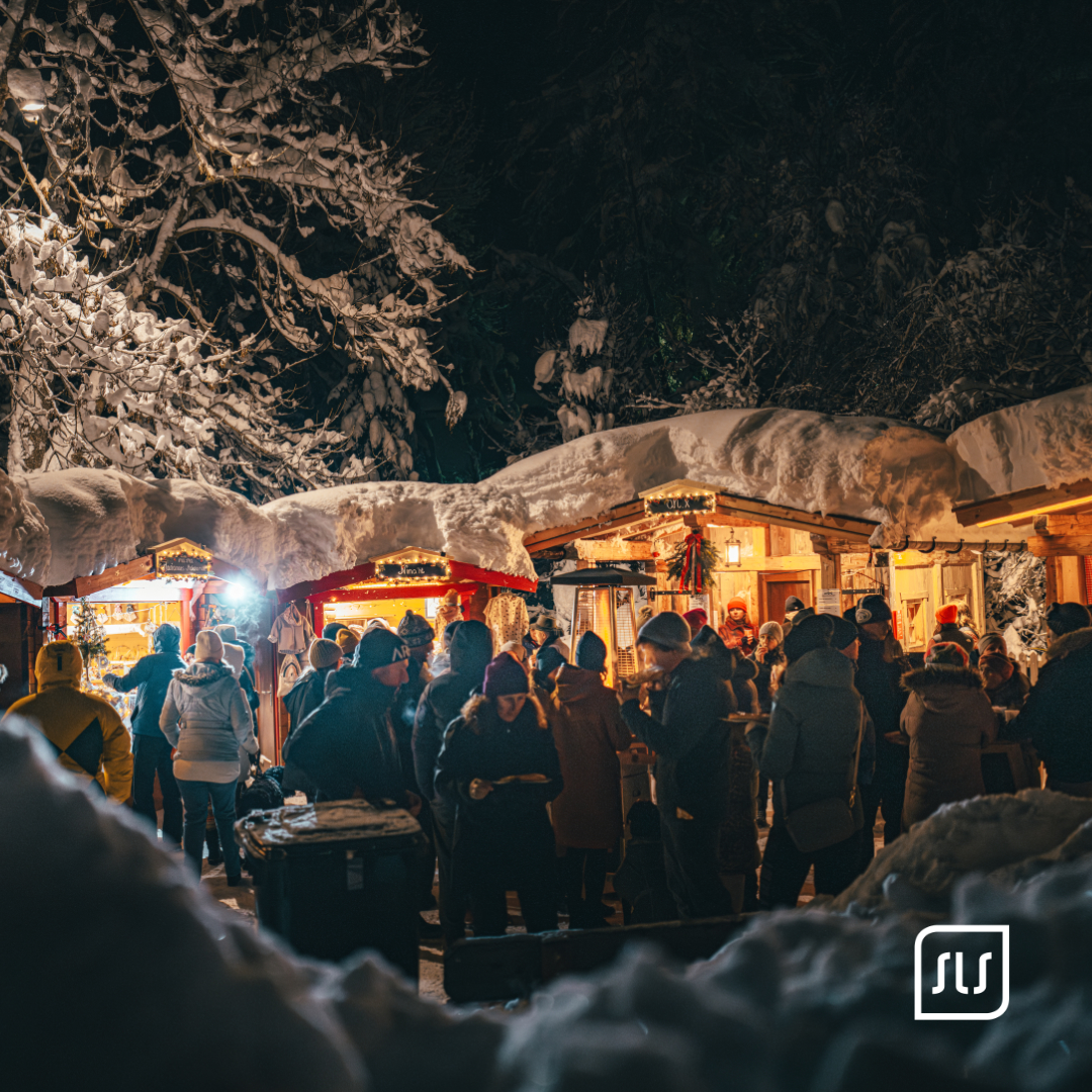 Besucher stehen abends zwischen beleuchteten, verschneiten Holzhütten beim Adventzauber in St. Anton