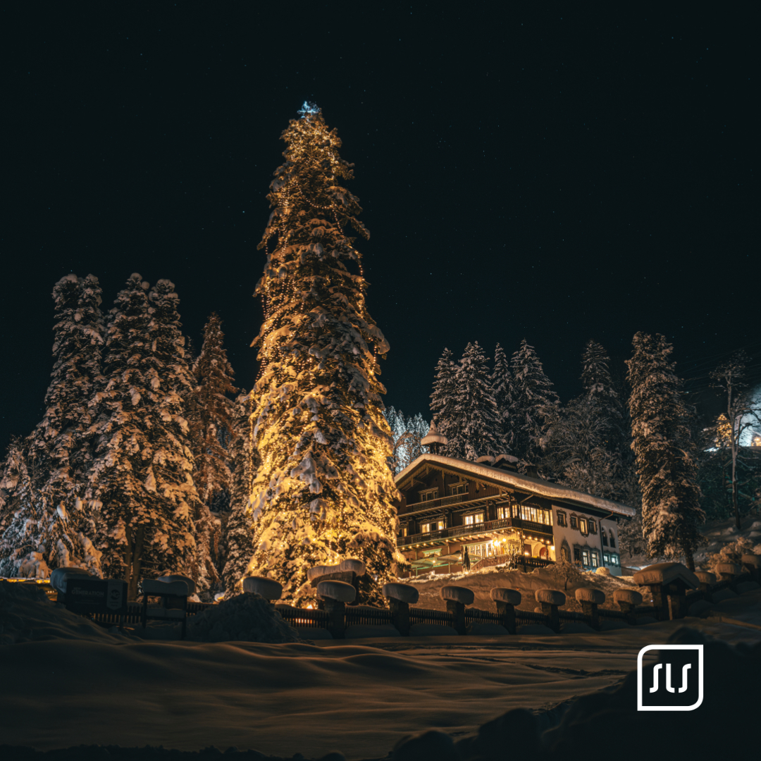 Großer schneebedeckter Baum wird nachts warm beleuchtet; dahinter steht ein Gebäude in St. Anton