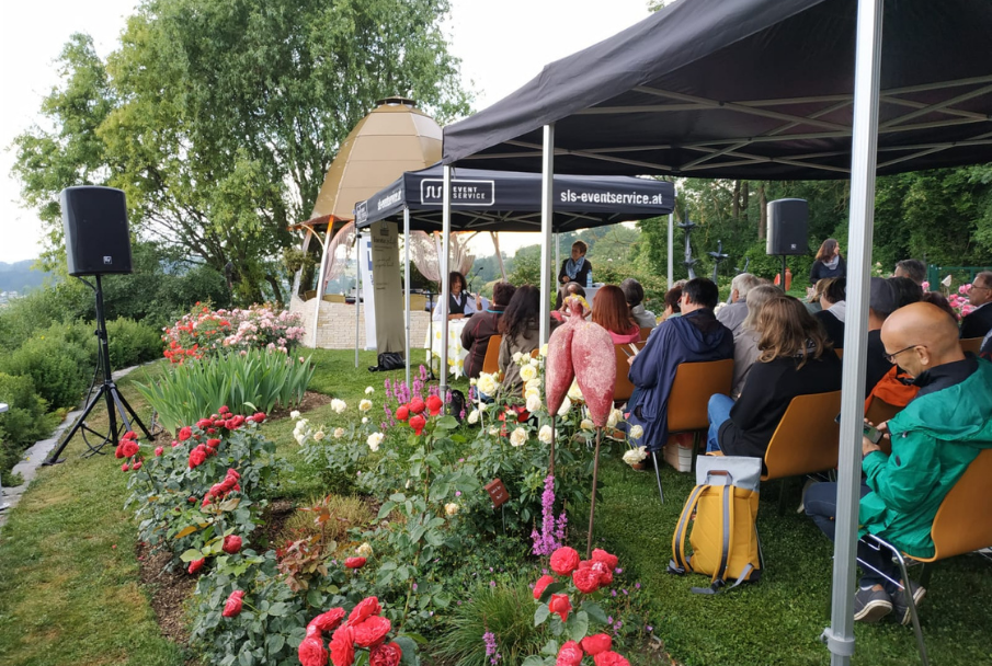 Publikum sitzt unter schwarzen SLS-Faltzelten bei einer Lesung neben Rosenbeeten im Freien