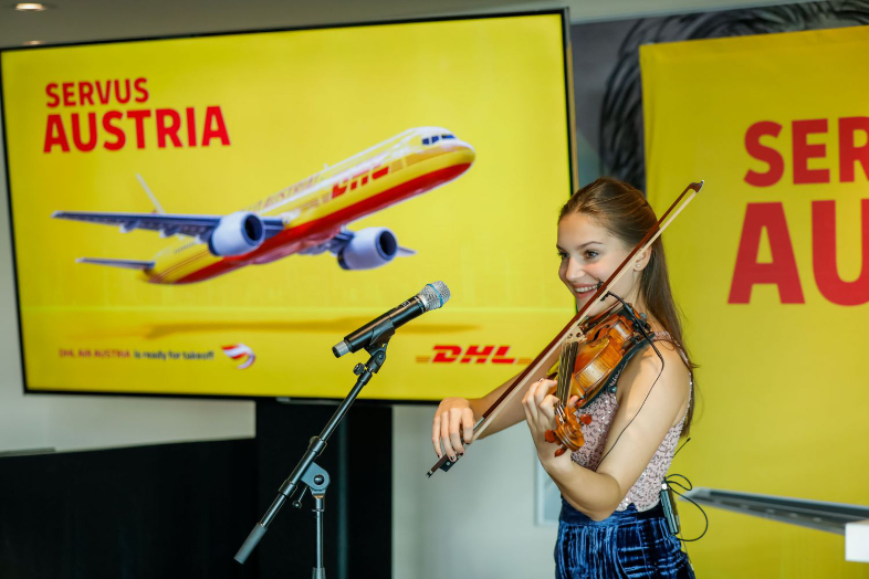 Violinistin am Mikrofon vor einer DHL-Präsentation mit Flugzeug und Schriftzug „Servus Austria“