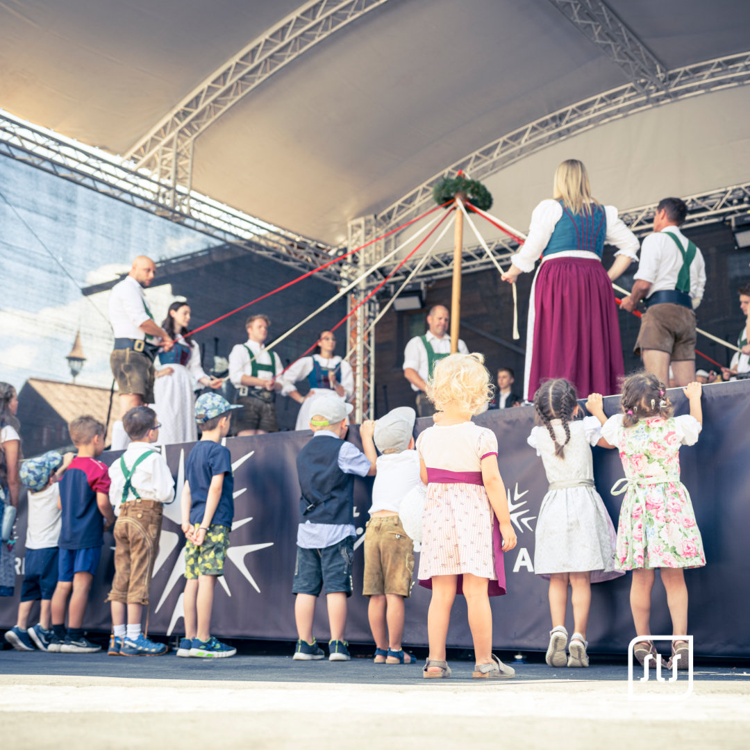 Erwachsene in Tracht halten bunte Bänder um einen geschmückten Pfosten auf der Dorffest-Bühne; Kinder sehen zu