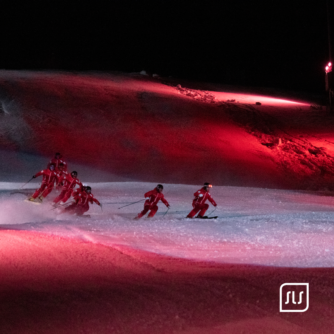 Gruppe von Skifahrern in roten Anzügen fährt bei der JWSC-Eröffnung 2023 in Formation über die beleuchtete Piste