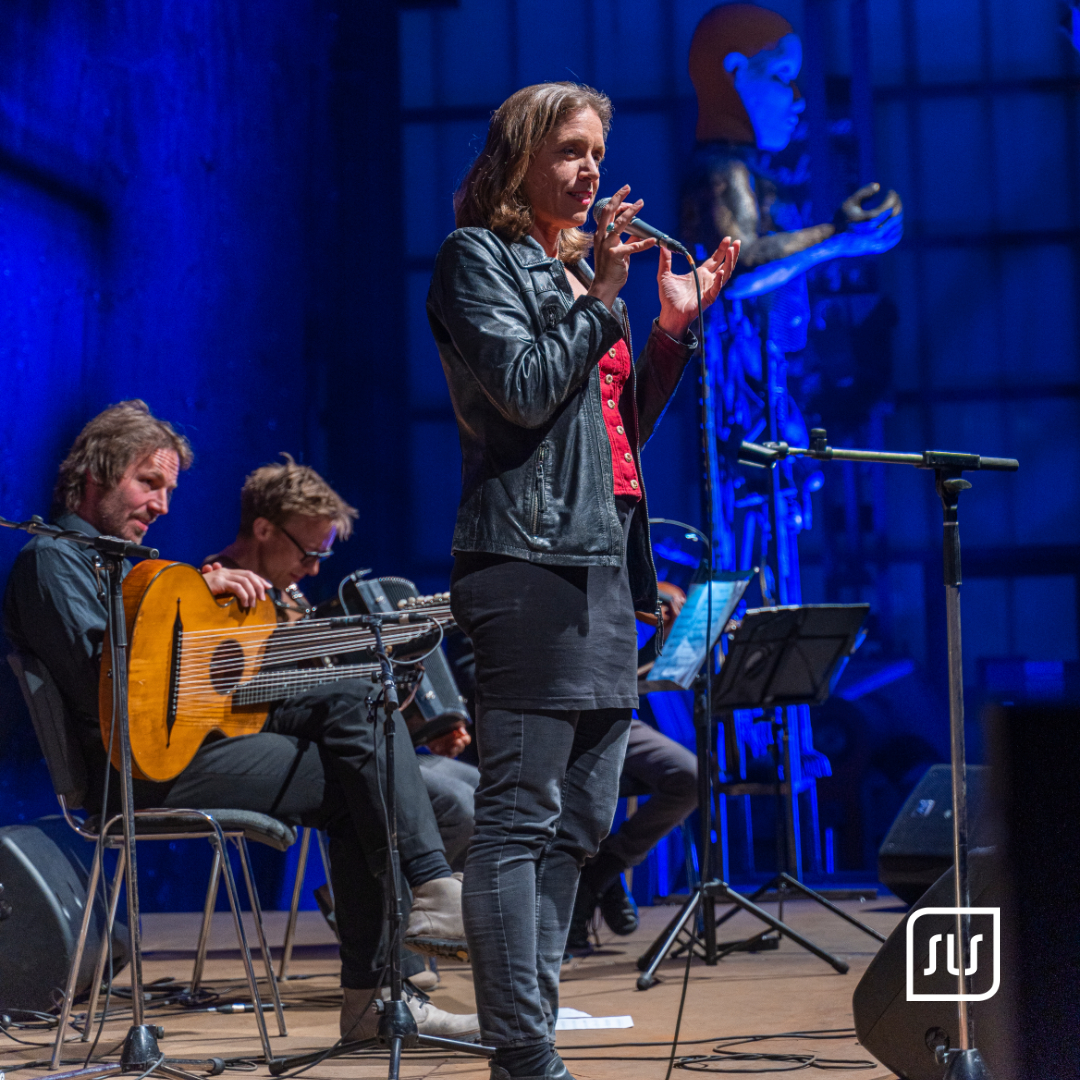 Agnes Palmisano steht mit Mikrofon vor sitzenden Musikern mit Gitarre auf einer blau beleuchteten Bühne
