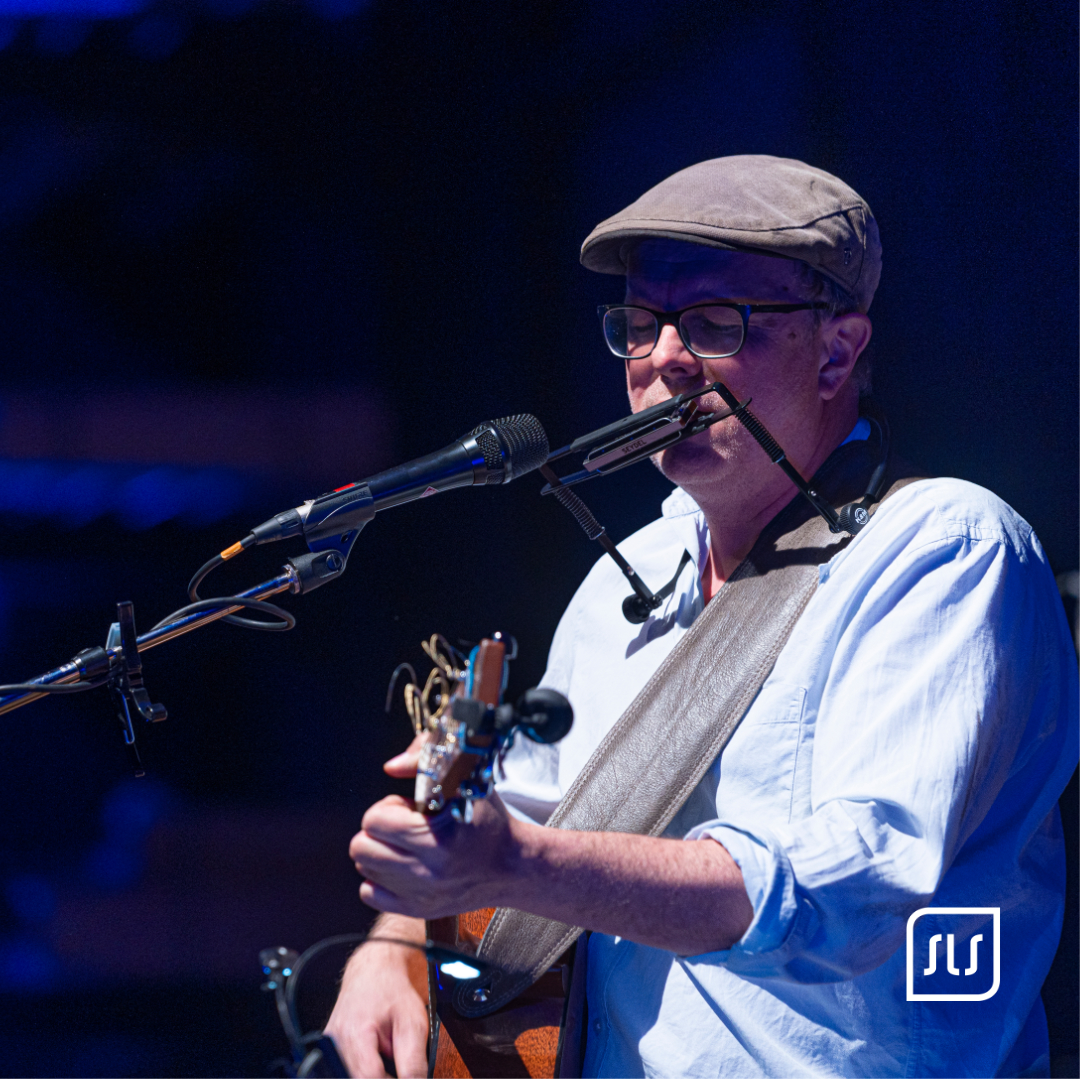 Musiker mit Schiebermütze spielt Gitarre und Mundharmonika vor einem Mikrofon auf blau beleuchteter Bühne