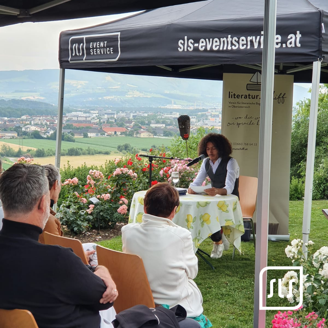 Vortragende Person liest an einem Tisch unter schwarzem SLS-Faltzelt vor sitzendem Publikum