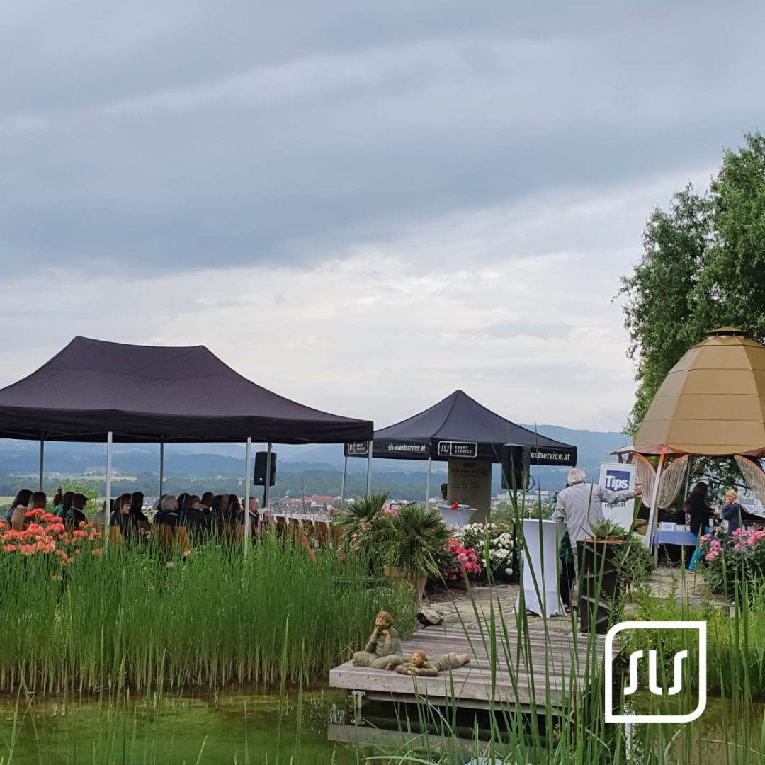 Dunkle Veranstaltungszelte mit Gästen neben einem Teich, Blumen und Schilf unter bewölktem Himmel