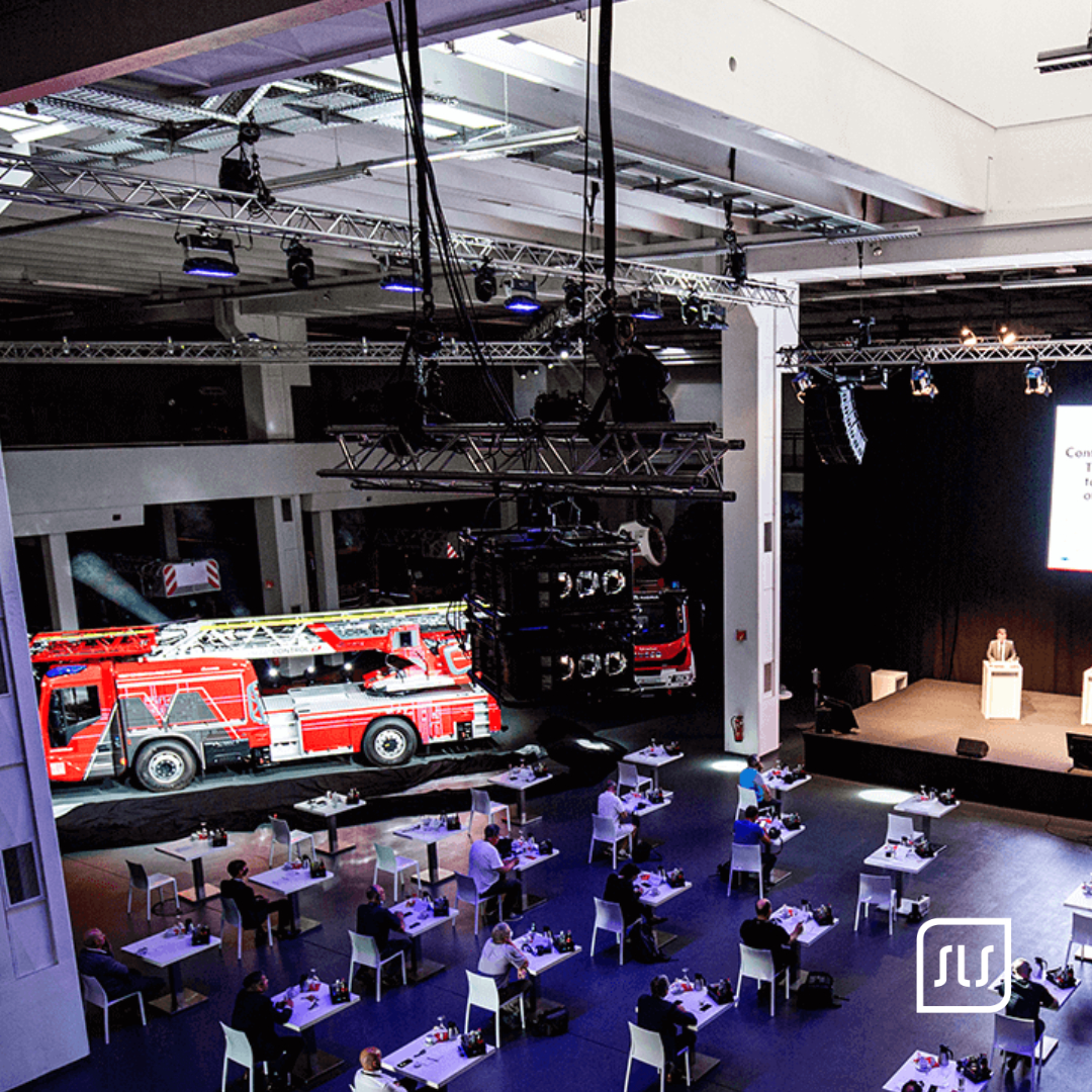 Blick von oben auf ein Magirus-Feuerwehrfahrzeug, die Bühne und Gäste an Einzeltischen in der Halle