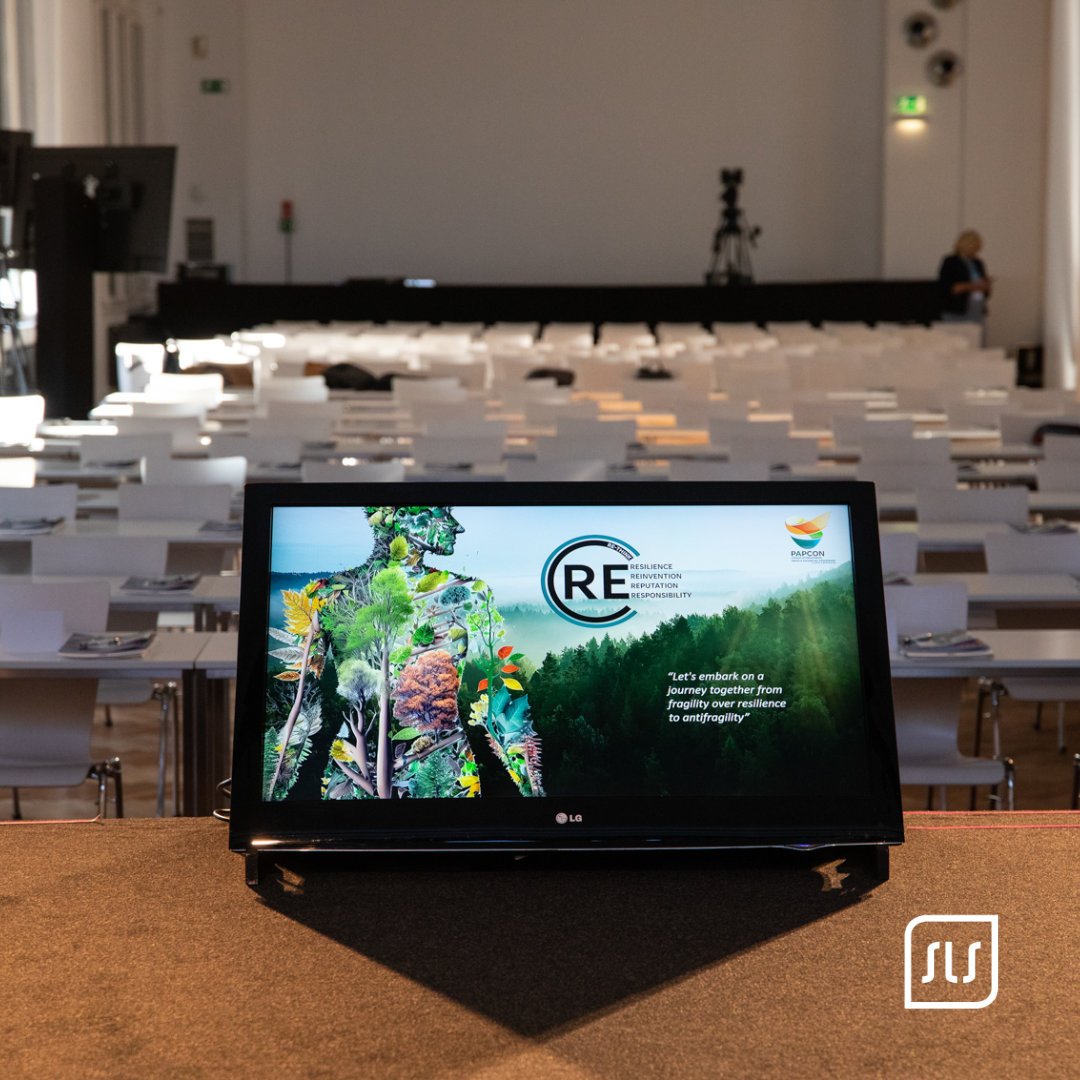 Monitor mit PAPCON-Präsentation steht vor Reihen weißer Konferenzstühle im noch leeren Saal