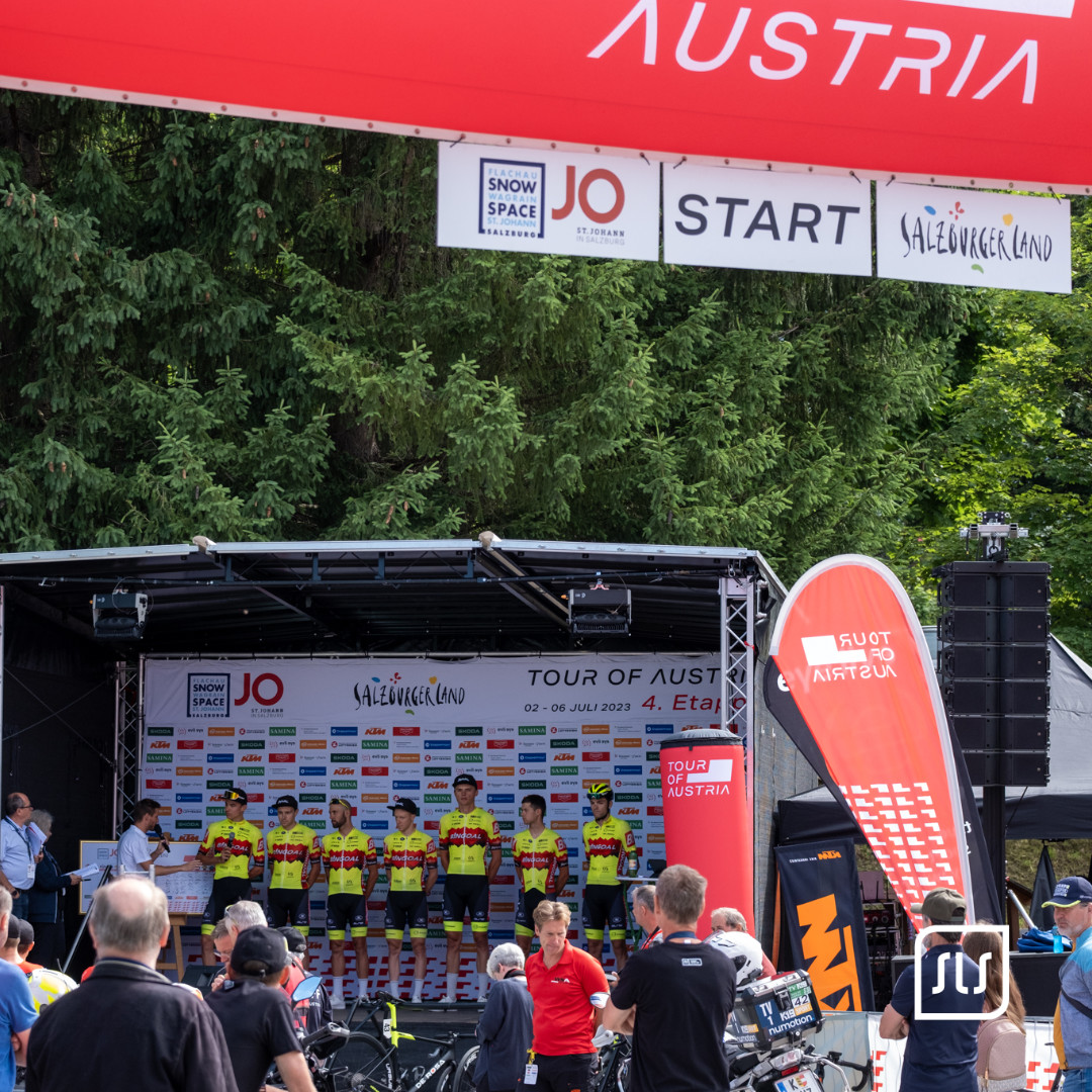 Radteam steht auf der Bühne unter dem Startbanner der Tour of Austria 2023 in St. Johann