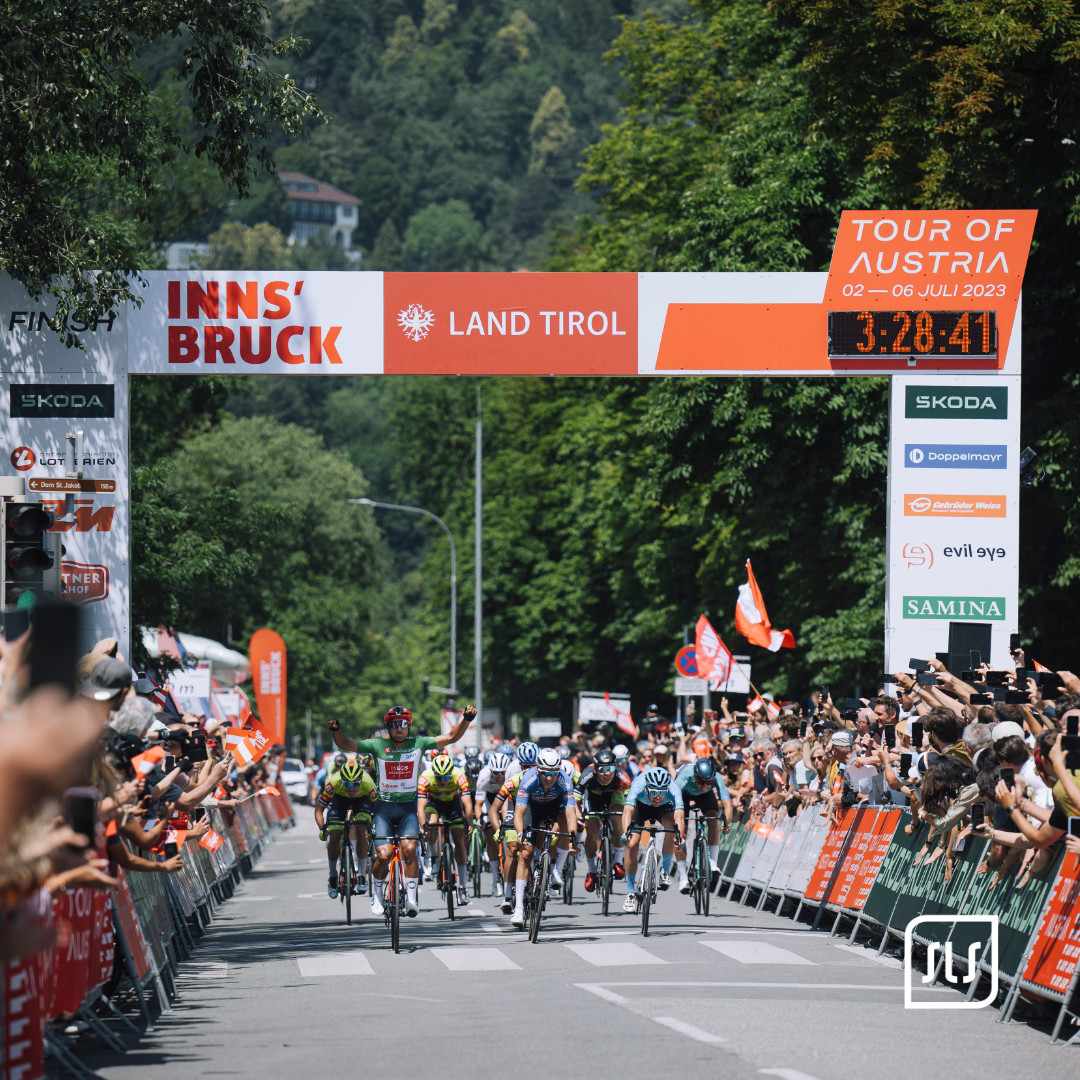 Rennradfahrer fahren unter dem Zielbogen der Tour of Austria 2023 in Innsbruck vor jubelndem Publikum