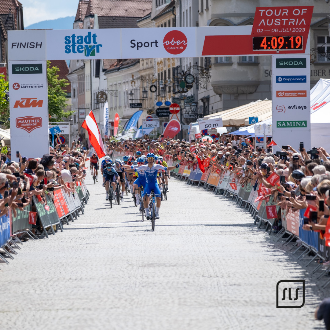 Rennradfahrer fahren unter dem Zielbogen der Tour of Austria 2023 in Steyr durch eine von Zuschauern gesäumte Straße