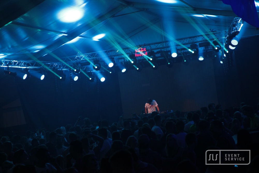 DJ auf einer Bühne vor großem Publikum mit türkiser und weißer Lichtshow