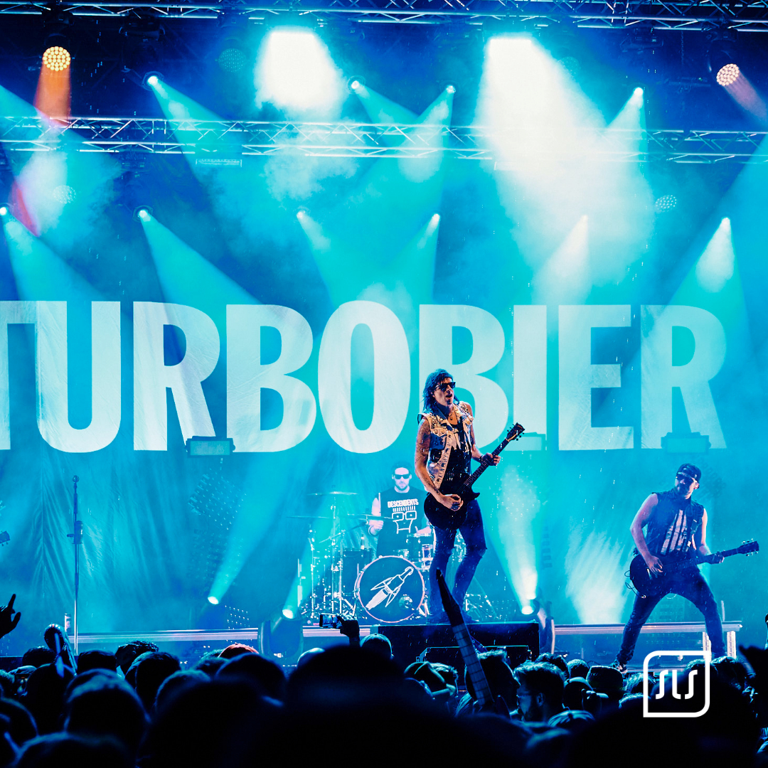 Band Turbobier mit Sänger, Gitarrist und Schlagzeuger auf blau beleuchteter Bühne vor Publikum
