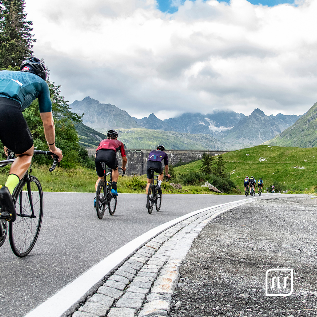 Mehrere Rennradfahrende fahren auf einer Kurve durch grüne Berglandschaft vor alpinen Gipfeln beim Arlberg Giro