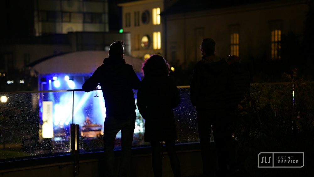 Drei Personen von hinten an einer Glasbrüstung mit Blick auf die blau beleuchtete Konzertbühne
