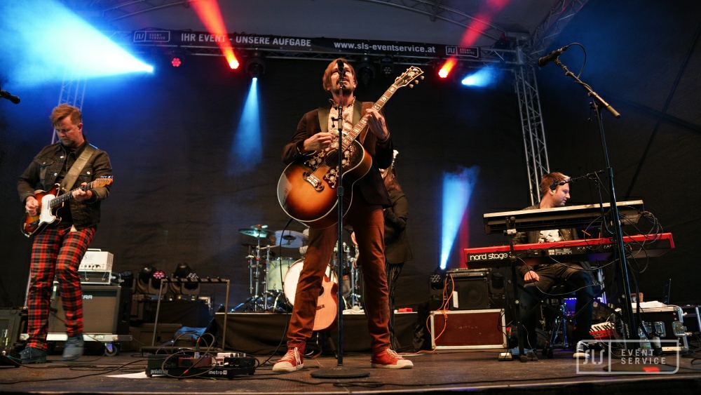 Drei Musiker auf der Konzertbühne mit E-Gitarre, Akustikgitarre und Keyboard unter blau-rotem Licht