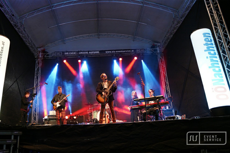 Weite Ansicht einer Liveband mit Gitarren, Keyboard und Schlagzeug auf einer blau-rot beleuchteten Bühne