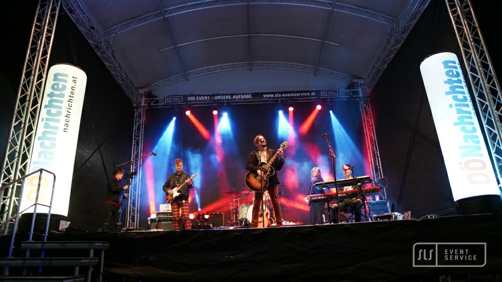 Liveband mit Sänger und Gitarre auf einer überdachten Bühne im blauen und roten Licht