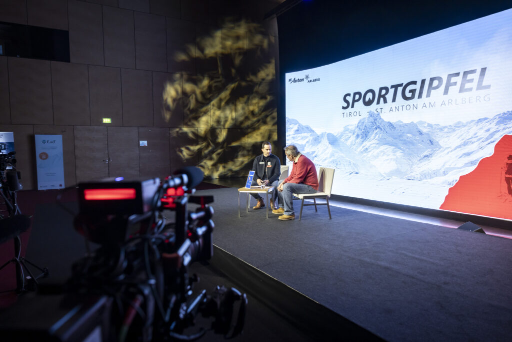 Zwei Gesprächspartner auf der Bühne des Sportgipfels Tirol vor einer LED-Wall mit Bergmotiv; Kamera im Vordergrund