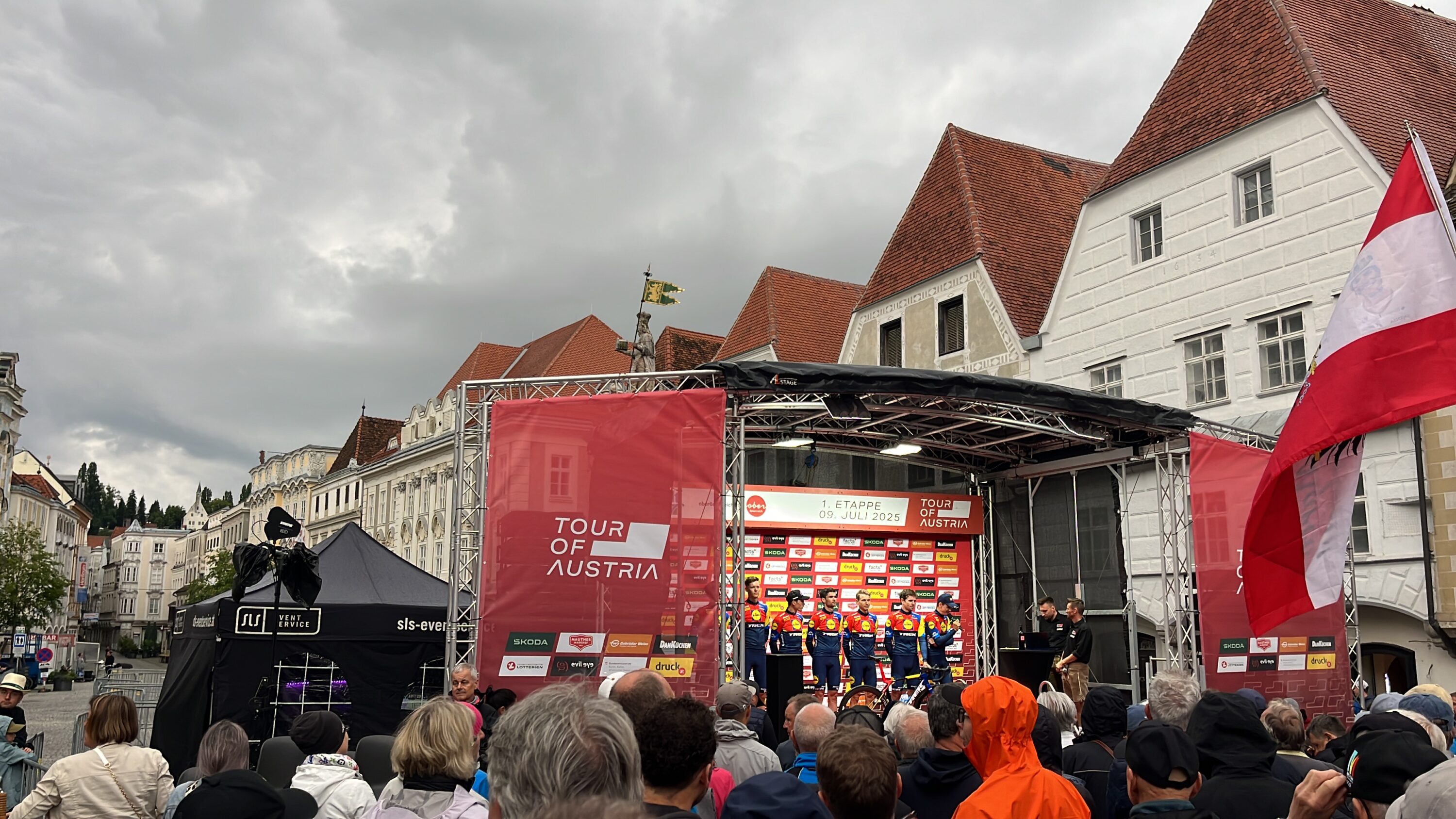 Radsportteam auf der überdachten Bühne der Tour of Austria 2025 mit roter Veranstaltungsbeschilderung