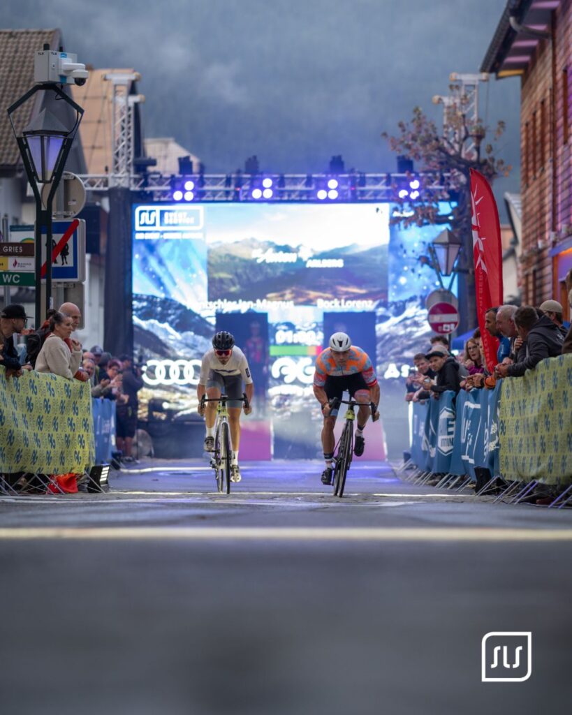 Zwei Radfahrer erreichen zwischen Absperrgittern und Zuschauern das beleuchtete Ziel des Arlberg Giro 2026